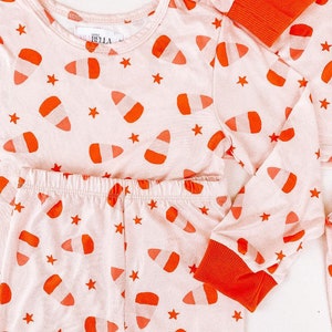 May include: Pink pajamas with long sleeves and pants. The pajamas have a candy corn print with orange and white candy corn on a pink background. The pajamas also have small white stars on the fabric. The pajamas have orange cuffs on the sleeves and legs.