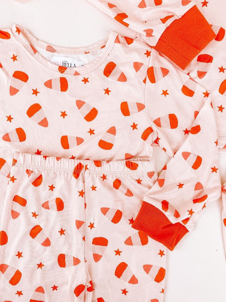 May include: Pink pajamas with long sleeves and pants. The pajamas have a candy corn print with orange and white candy corn on a pink background. The pajamas also have small white stars on the fabric. The pajamas have orange cuffs on the sleeves and legs.
