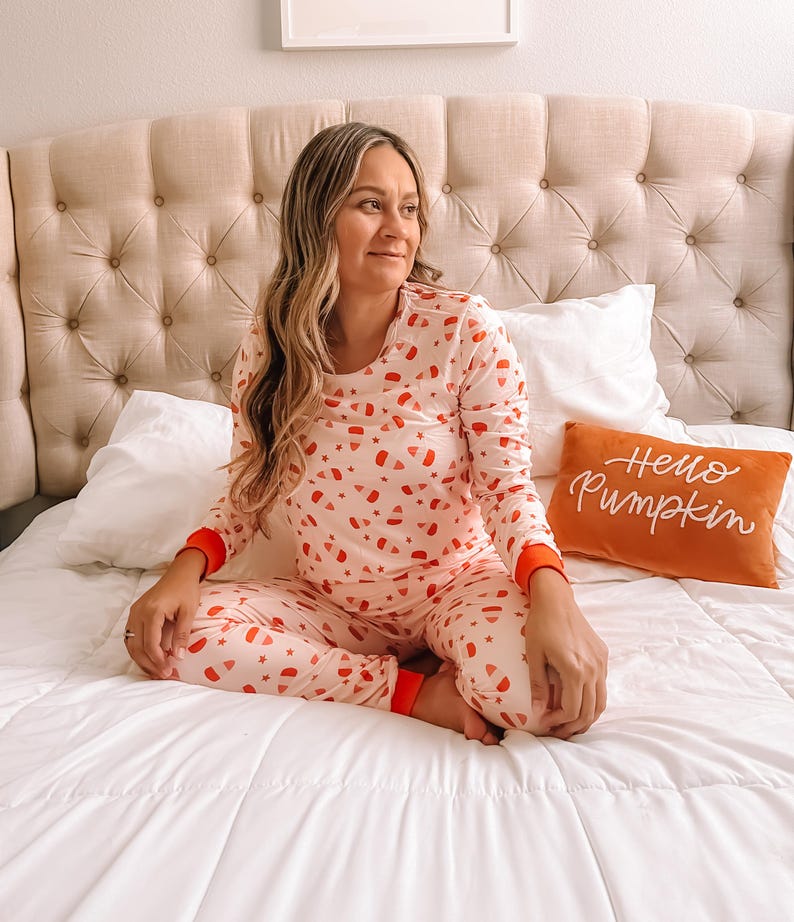 May include: A woman wearing pink pajamas with a candy corn print sits on a white bed with a pillow that says "Hello Pumpkin".