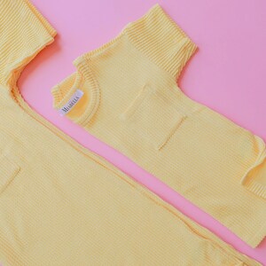 May include: Two-piece yellow waffle-knit outfit, possibly pajamas or loungewear, with a pocket on the top and a label that reads "MABELLA". The items are laid out on a pink background.