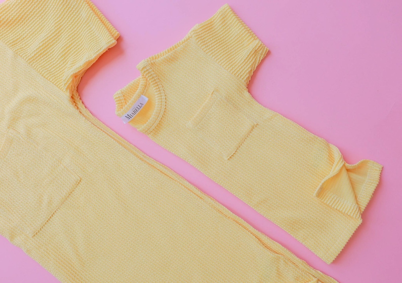 May include: Two-piece yellow waffle-knit outfit, possibly pajamas or loungewear, with a pocket on the top and a label that reads "MABELLA". The items are laid out on a pink background.