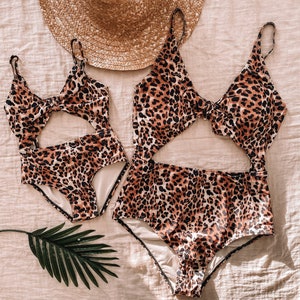 Pu&ograve; includere: Due costumi da bagno con stampa leopardata, uno per adulti e uno per bambini, con dettagli ritagliati. Un cappello di paglia &egrave; posto sopra i costumi da bagno su una superficie dai toni neutri. Una foglia di palma verde &egrave; posizionata vicino ai costumi da bagno.