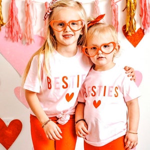 Pode incluir: Duas meninas pequenas vestindo camisas rosa e brancas com o texto "BESTIE BESTIES" e detalhes de cora&ccedil;&atilde;o vermelho. Ambas usam &oacute;culos vermelhos em forma de cora&ccedil;&atilde;o e t&ecirc;m cabelo loiro. As meninas est&atilde;o em p&eacute; em frente a um fundo de cora&ccedil;&otilde;es rosa e vermelhos.