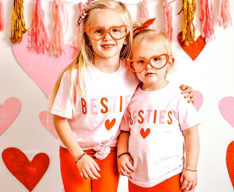 Pode incluir: Duas meninas pequenas vestindo camisas rosa e brancas com o texto "BESTIE BESTIES" e detalhes de cora&ccedil;&atilde;o vermelho. Ambas usam &oacute;culos vermelhos em forma de cora&ccedil;&atilde;o e t&ecirc;m cabelo loiro. As meninas est&atilde;o em p&eacute; em frente a um fundo de cora&ccedil;&otilde;es rosa e vermelhos.