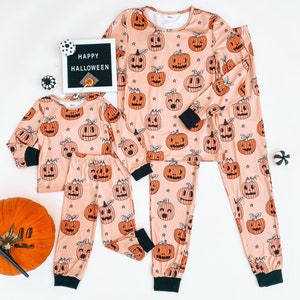 May include: A set of matching pajamas featuring a peach-colored background with a repeating pattern of orange jack-o'-lantern faces. The pajamas come in two sizes, one for an adult and one for a child. The text "HAPPY HALLOWEEN" is visible on a black letter board in the background.