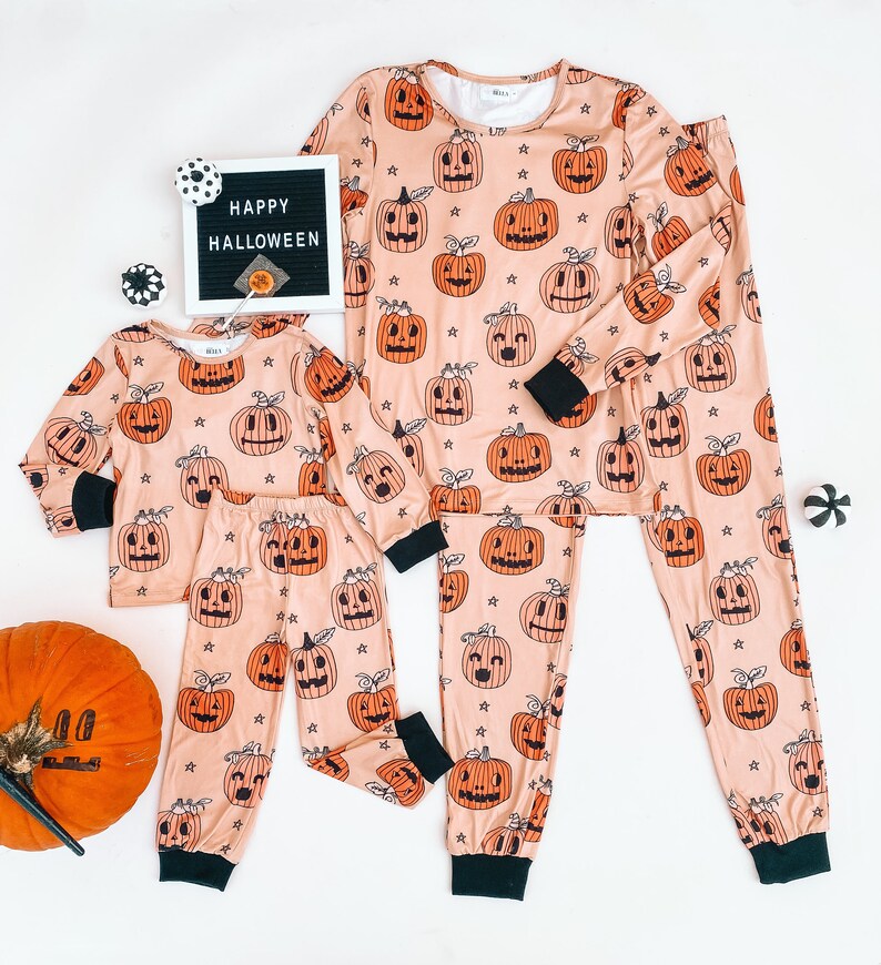 May include: A set of matching pajamas featuring a peach-colored background with a repeating pattern of orange jack-o'-lantern faces. The pajamas come in two sizes, one for an adult and one for a child. The text "HAPPY HALLOWEEN" is visible on a black letter board in the background.