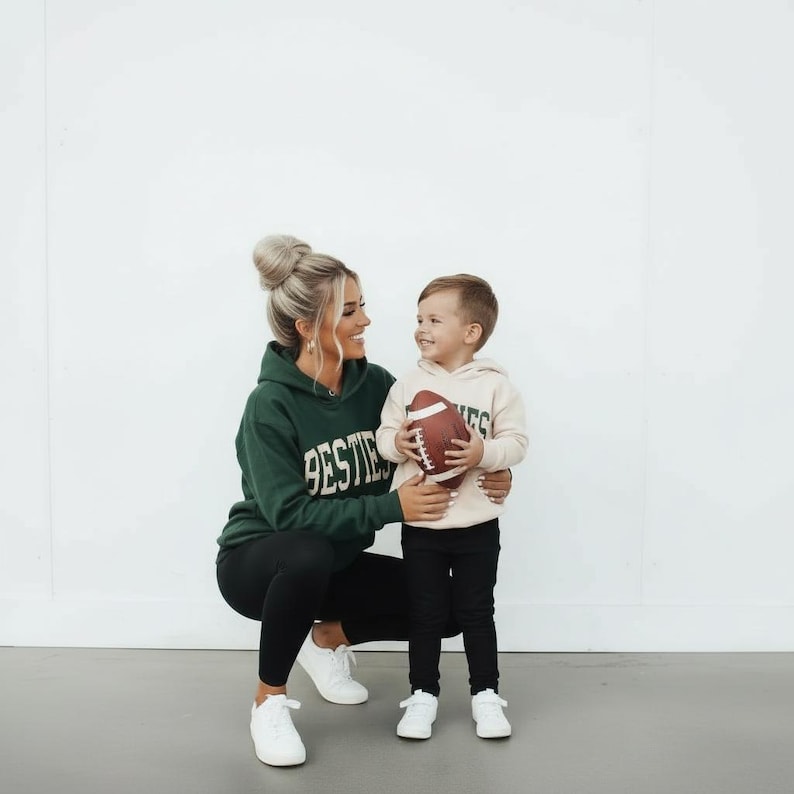 May include: A woman and a child wearing matching hoodies with the word "BESTIES" printed on them. The woman is wearing a dark green hoodie and black leggings, while the child is wearing a beige hoodie and black pants. The child is holding a brown football.