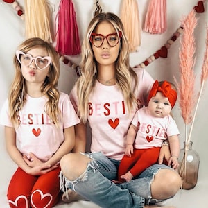 Pode incluir: Tr&ecirc;s pessoas vestindo camisetas rosa com a palavra "Bestie" e um cora&ccedil;&atilde;o vermelho na frente. As camisetas s&atilde;o usadas por uma mulher com cabelo longo e loiro e &oacute;culos em forma de cora&ccedil;&atilde;o, uma menina com cabelo longo e castanho e &oacute;culos em forma de cora&ccedil;&atilde;o, e uma menina beb&ecirc; com uma faixa de cabe&ccedil;a vermelha. A mulher est&aacute; usando jeans azul, a menina est&aacute; usando cal&ccedil;as vermelhas, e a menina beb&ecirc; est&aacute; usando cal&ccedil;as vermelhas.