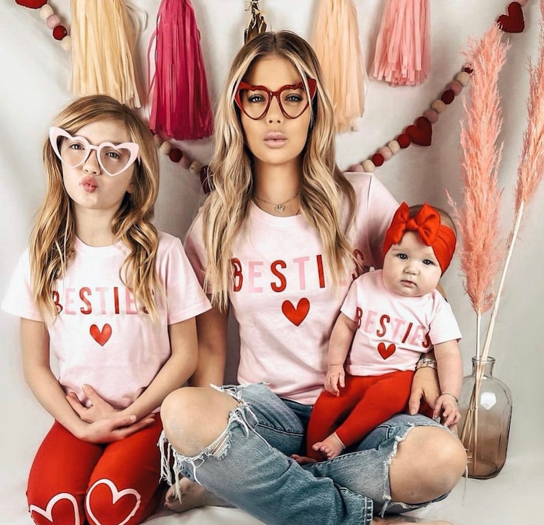 Pode incluir: Tr&ecirc;s pessoas vestindo camisetas rosa com a palavra "Bestie" e um cora&ccedil;&atilde;o vermelho na frente. As camisetas s&atilde;o usadas por uma mulher com cabelo longo e loiro e &oacute;culos em forma de cora&ccedil;&atilde;o, uma menina com cabelo longo e castanho e &oacute;culos em forma de cora&ccedil;&atilde;o, e uma menina beb&ecirc; com uma faixa de cabe&ccedil;a vermelha. A mulher est&aacute; usando jeans azul, a menina est&aacute; usando cal&ccedil;as vermelhas, e a menina beb&ecirc; est&aacute; usando cal&ccedil;as vermelhas.