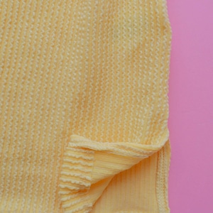May include: Close-up of a textured, pale yellow fabric with a ribbed pattern. The fabric is folded at the bottom, revealing a smooth, solid yellow underside. The background is a solid pink.