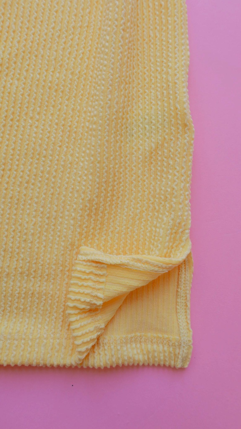 May include: Close-up of a textured, pale yellow fabric with a ribbed pattern. The fabric is folded at the bottom, revealing a smooth, solid yellow underside. The background is a solid pink.