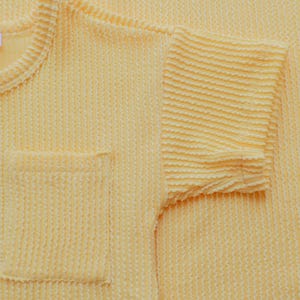 May include: Close-up of a light yellow textured knit shirt with a pocket on the front. The short sleeve is visible, and the fabric has a ribbed pattern. The shirt is made of a soft, comfortable material.