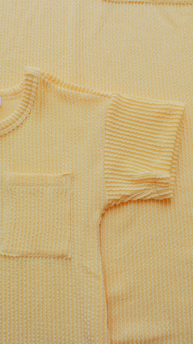 May include: Close-up of a light yellow textured knit shirt with a pocket on the front. The short sleeve is visible, and the fabric has a ribbed pattern. The shirt is made of a soft, comfortable material.