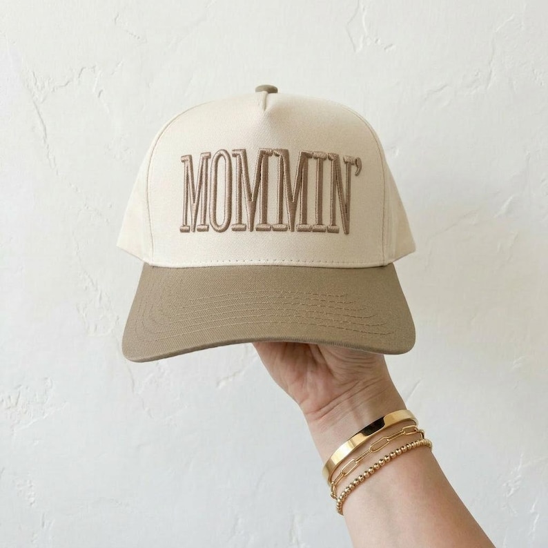 May include: A beige and tan baseball cap with the word "MOMMIN" embroidered in brown. The cap has a curved brim and a structured crown. The hat is a casual accessory.