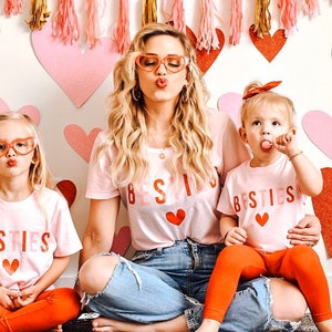 Pode incluir: Tr&ecirc;s pessoas vestindo camisetas rosa com a palavra "Besties" impressa. As camisetas t&ecirc;m um design de cora&ccedil;&atilde;o vermelho. As pessoas posam em frente a uma parede branca decorada com cora&ccedil;&otilde;es rosas.