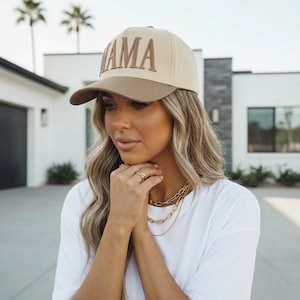 May include: A beige and brown baseball cap with the word "MAMA" embroidered on the front. The hat has a curved brim and a structured fit. The cap is a stylish accessory.