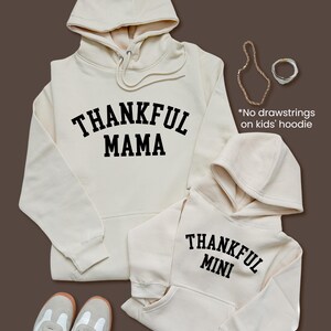 May include: Two cream-colored hoodies with the words "THANKFUL MAMA" and "THANKFUL MINI" printed in black. The hoodies have drawstrings and a front pocket. A pair of sneakers and jewelry are also in the image.