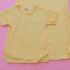 May include: A light yellow textured short-sleeved shirt with a chest pocket and a matching dress. The shirt has a small tag that reads "MIABELLA". The garments are made of a ribbed fabric and are set against a pink background.
