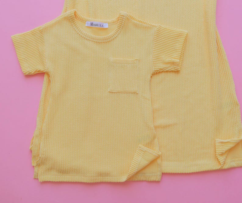 May include: A light yellow textured short-sleeved shirt with a chest pocket and a matching dress. The shirt has a small tag that reads "MIABELLA". The garments are made of a ribbed fabric and are set against a pink background.