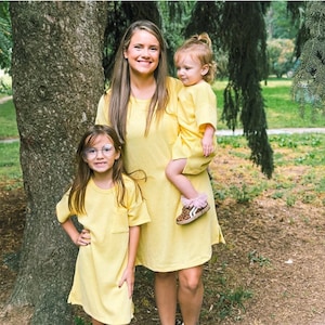 May include: A family of three wearing matching yellow dresses. The dresses are short-sleeved and have a simple design. The youngest child wears leopard print shoes with pink trim. The family is standing in a park setting with a large tree in the background.