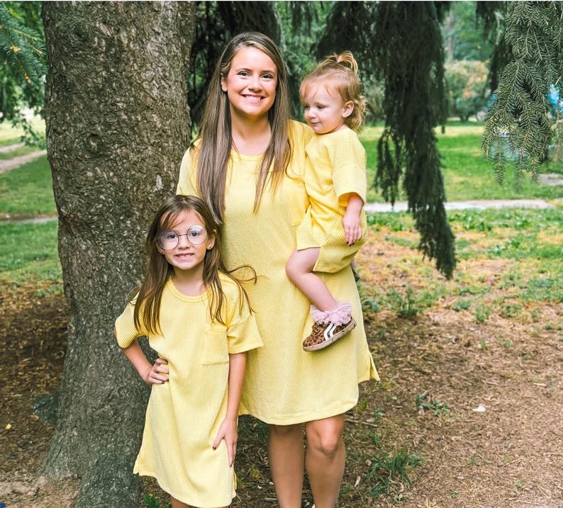 May include: A family of three wearing matching yellow dresses. The dresses are short-sleeved and have a simple design. The youngest child wears leopard print shoes with pink trim. The family is standing in a park setting with a large tree in the background.