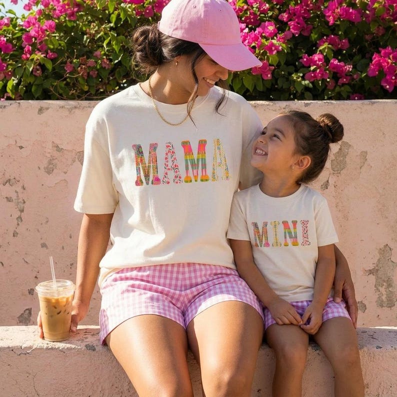 May include: Two cream-coloured t-shirts with the words "MAMA" and "MINI" in a colourful floral print. The mother wears a pink baseball cap and pink gingham shorts. The child wears matching shorts. A clear plastic cup with a straw is visible.