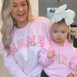 May include: A pink sweatshirt with the word "MAMA" in gold glitter lettering. A smaller pink sweatshirt with the word "MINI" in gold glitter lettering is worn by a child.