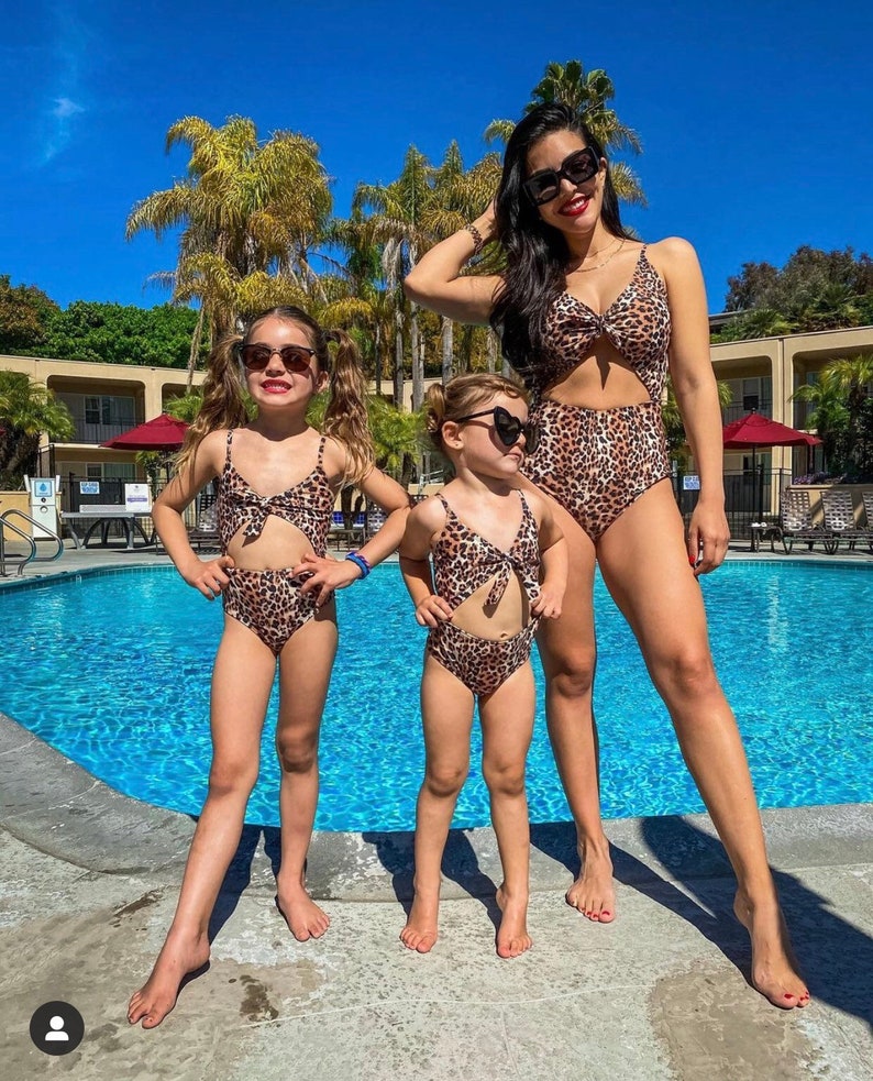 Pu&ograve; includere: Tre persone con costumi da bagno leopardati abbinati e occhiali da sole posano vicino a una piscina. I costumi da bagno includono un costume intero e costumi da bagno a due pezzi. Palme e cielo blu sullo sfondo.