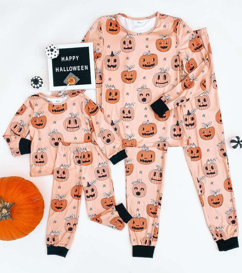 May include: A set of matching pajamas featuring a peach background with a repeating pattern of orange jack-o'-lantern faces. The pajamas come in two sizes, one for an adult and one for a child. The text "HAPPY HALLOWEEN" is visible on a black letter board in the background.