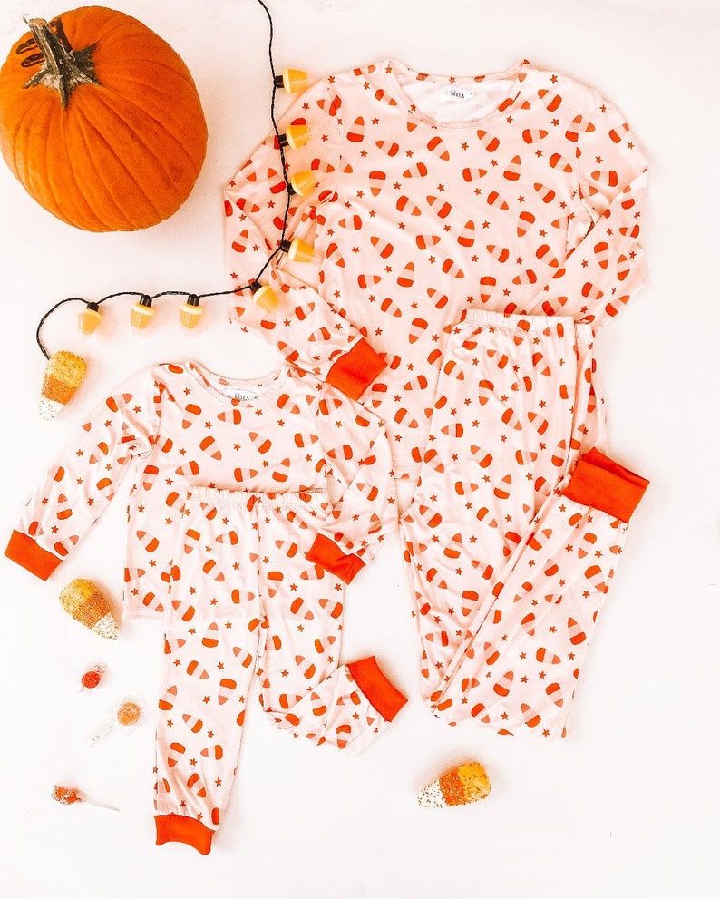 May include: A set of two pairs of pajamas with a candy corn print. The pajamas are pink with orange and white candy corn designs. The pajamas are on a white background.