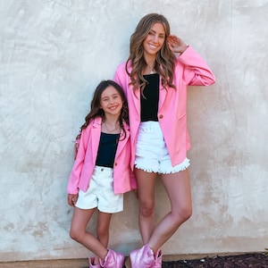 May include: Two people, a woman and a girl, wearing pink blazers and white denim shorts. They are both wearing pink sparkly cowboy boots and standing in front of a textured wall.