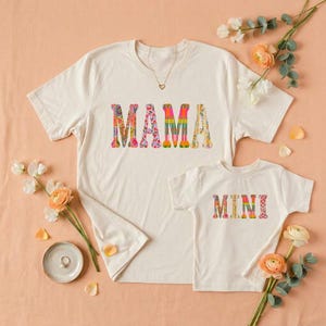 May include: Two cream-coloured t-shirts, one adult and one child size, with the words "MAMA" and "MINI" in colourful, patterned letters. The shirts feature floral and geometric designs. The image is styled with flowers and a necklace.