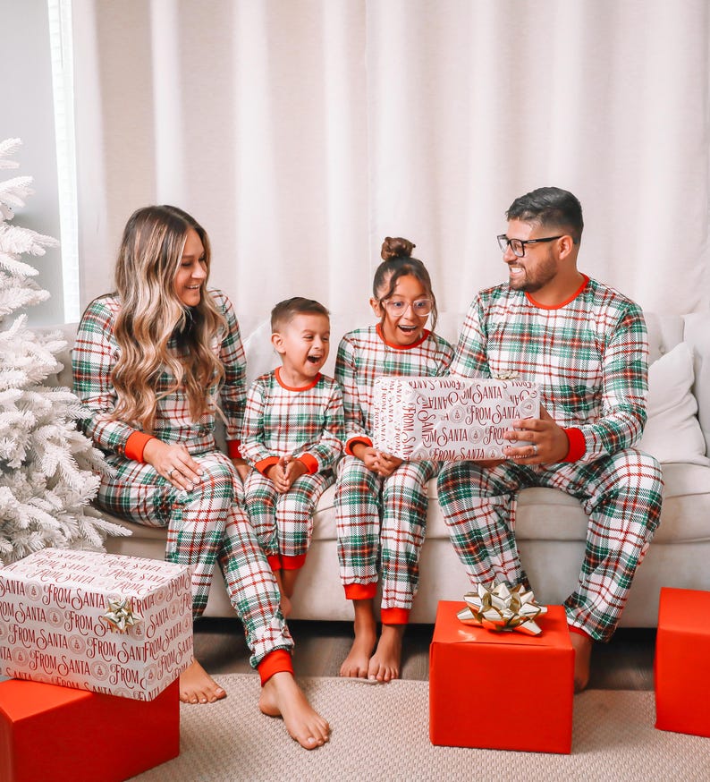 May include: Family matching Christmas pajamas. Red and green plaid print pajamas with white background.  Adults and children's sizes shown.  'From Santa' text is printed on gift wrap and pajamas.  A family is opening presents in front of a Christmas tree.