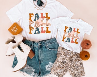 May include: Two white t-shirts with the word "FALL" printed in a retro style with a lightning bolt. The larger shirt has a sign that says "hello fall" in front of it. The smaller shirt is on top of leopard print shorts.