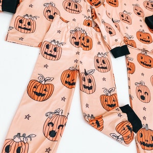 May include: A peach-colored pajama set with a long-sleeved top and pants. The pajamas feature a repeating pattern of orange jack-o'-lantern faces with black eyes and mouths. The background is a light peach color with small white stars.