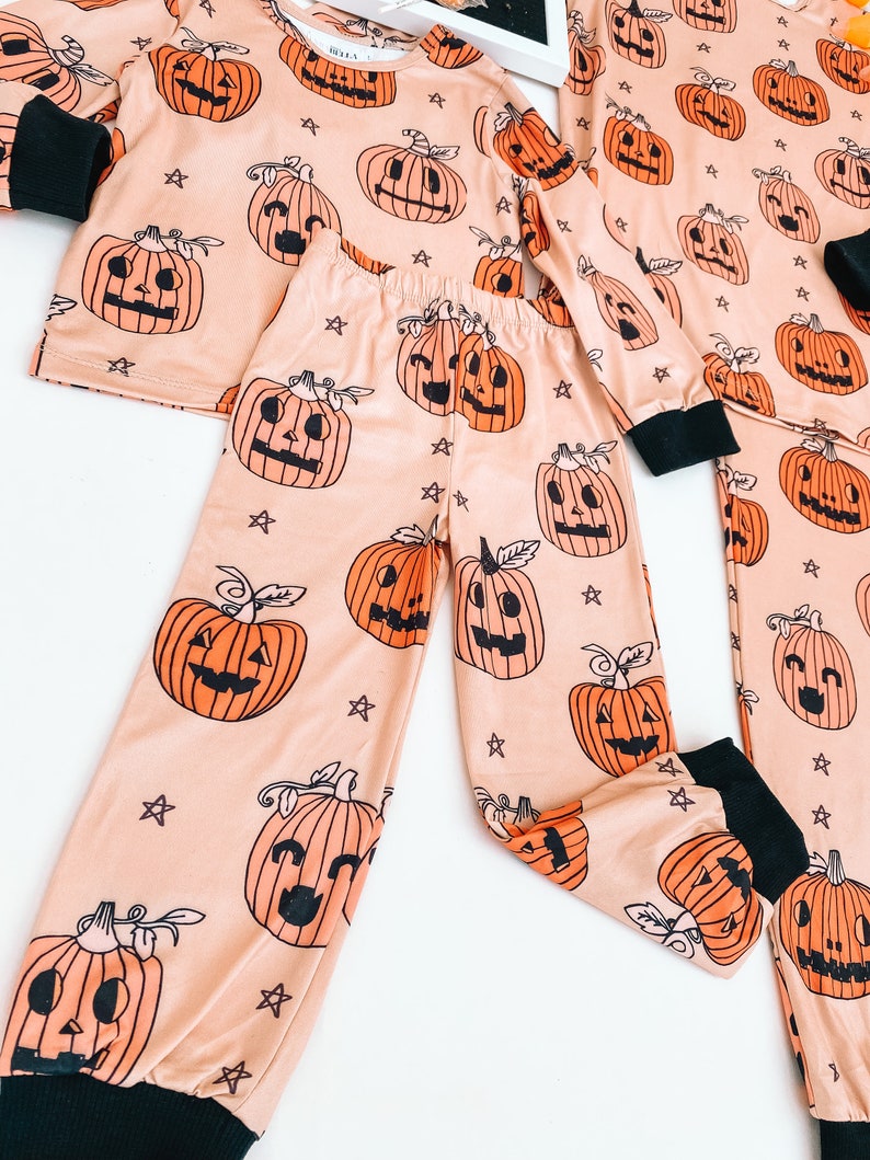 May include: A peach-colored pajama set with a long-sleeved top and pants. The pajamas feature a repeating pattern of orange jack-o'-lantern faces with black eyes and mouths. The background is a light peach color with small white stars.