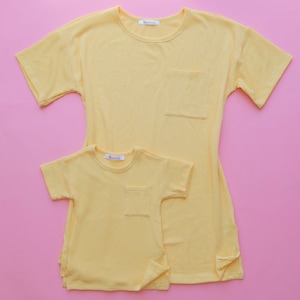 May include: Two yellow ribbed t-shirts, one adult and one child size, are displayed on a pink background. Each shirt has short sleeves, a chest pocket, and a small tag at the neckline. The adult shirt is longer than the child's.