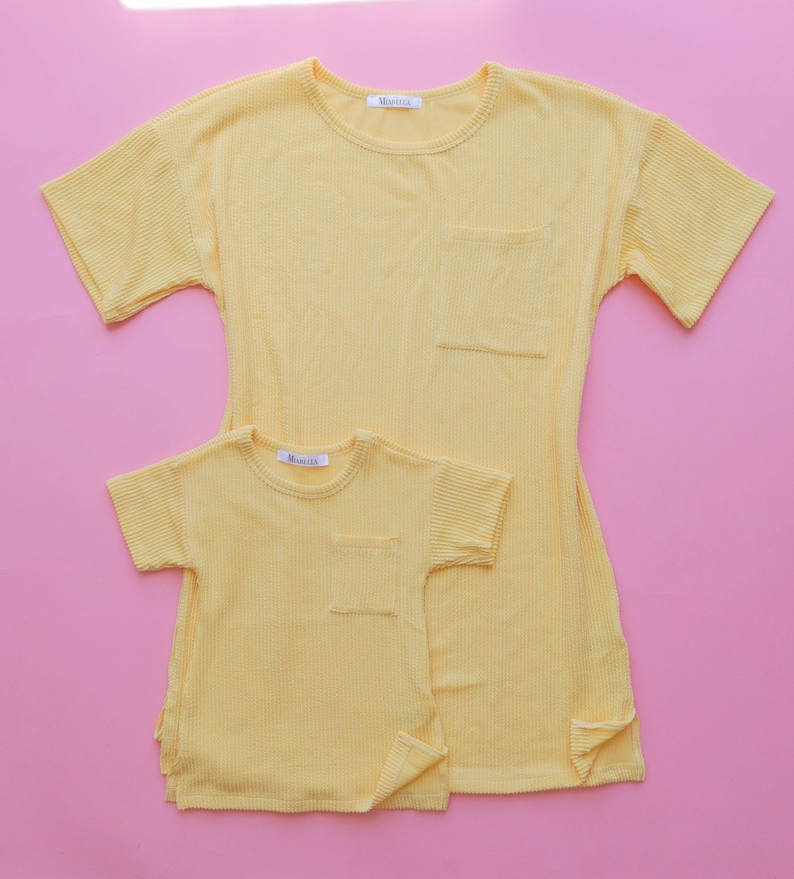 May include: Two yellow ribbed t-shirts, one adult and one child size, are displayed on a pink background. Each shirt has short sleeves, a chest pocket, and a small tag at the neckline. The adult shirt is longer than the child's.