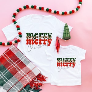 May include: Two white t-shirts with the text "merry merry Christmas" in green, red, and leopard print. The shirts are on a pink background with a red and white plaid blanket and a string of white and green pom-poms.