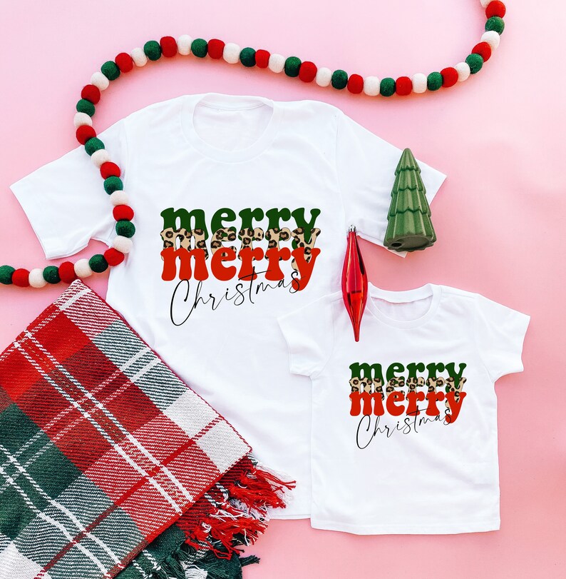 May include: Two white t-shirts with the text "merry merry Christmas" in green, red, and leopard print. The shirts are on a pink background with a red and white plaid blanket and a string of white and green pom-poms.