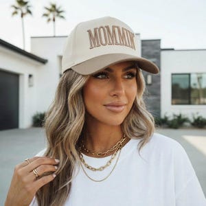 May include: A beige and brown baseball cap with the word "MOMMIN" embroidered on the front. The cap has a curved brim and is worn by a person. The person is wearing a white t-shirt and gold necklaces.