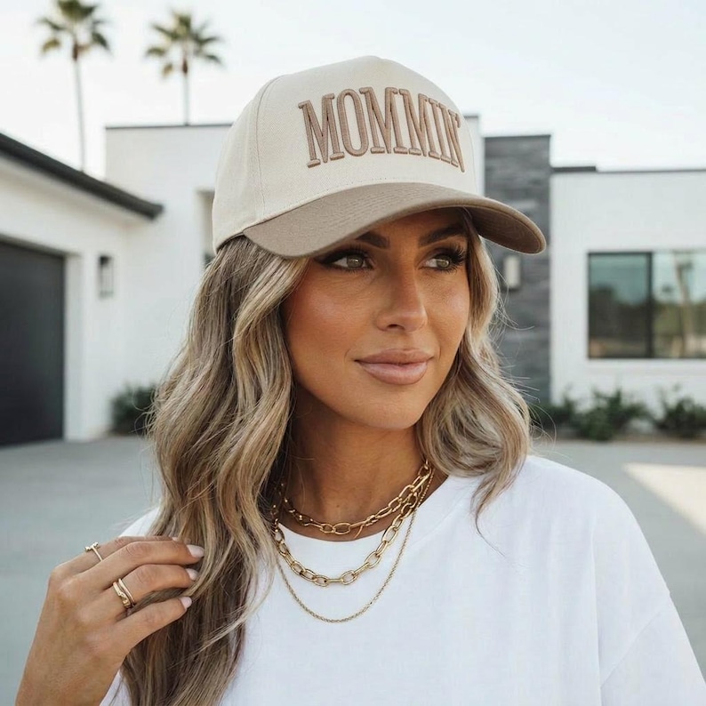 May include: A beige and brown baseball cap with the word "MOMMIN" embroidered on the front. The cap has a curved brim and is worn by a person. The person is wearing a white t-shirt and gold necklaces.