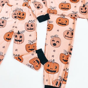 May include: A pair of matching pajamas with a peach background and black jack-o'-lantern print. The pajamas have black cuffs and a black waistband.