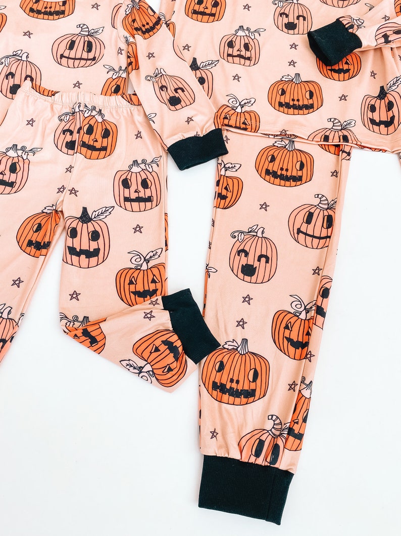 May include: A pair of matching pajamas with a peach background and black jack-o'-lantern print. The pajamas have black cuffs and a black waistband.