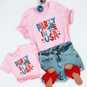 May include: Two pink t-shirts with the text "Party in the USA" in red, blue, and white. The shirts have a lightning bolt and stars. The shirts are on a white background with a pair of red, white, and blue sunglasses.