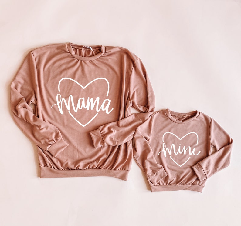 May include: Two long-sleeved shirts in a dusty rose color. The larger shirt has "mama" written in a white cursive font inside a white heart. The smaller shirt has "mini" written in a white cursive font inside a white heart.