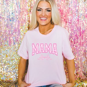 May include: A pink t-shirt with the word "MAMA" in pink letters with a white outline and a pink bow underneath. The shirt is being worn by a person in front of a pink sparkly background.