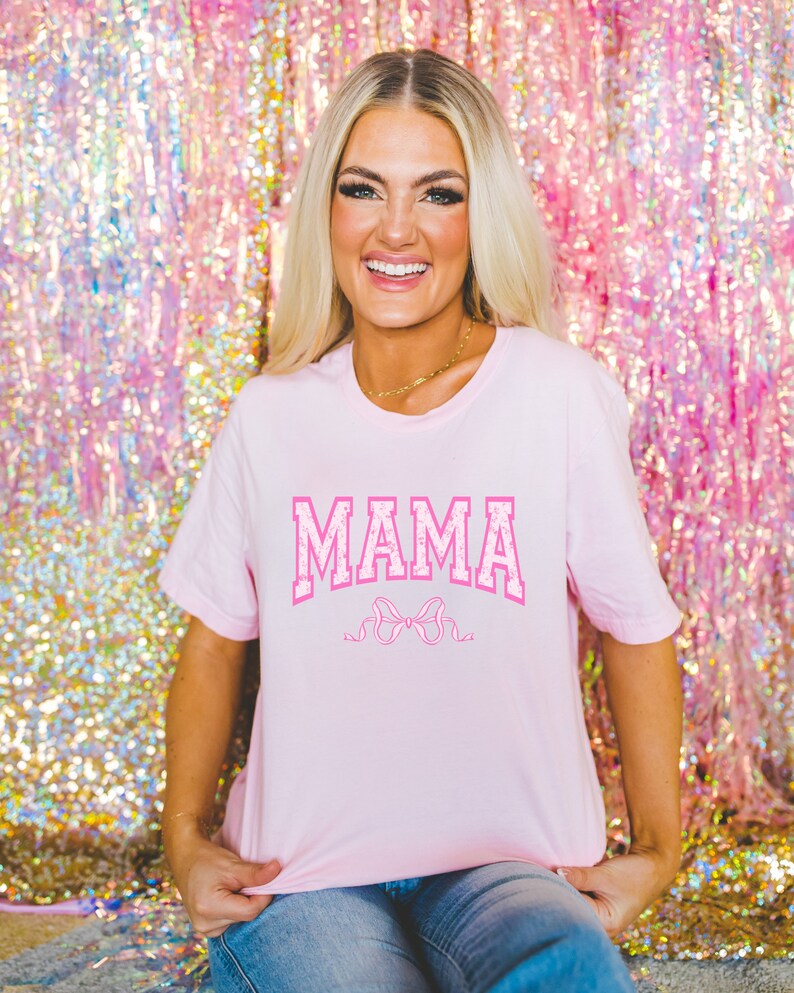 May include: A pink t-shirt with the word "MAMA" in pink letters with a white outline and a pink bow underneath. The shirt is being worn by a person in front of a pink sparkly background.