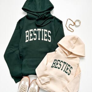 May include: Two hooded sweatshirts, one dark green and one beige, both with the word "BESTIES" in bold, white and green lettering. A pair of beige and white sneakers and gold jewelry are also in the image.