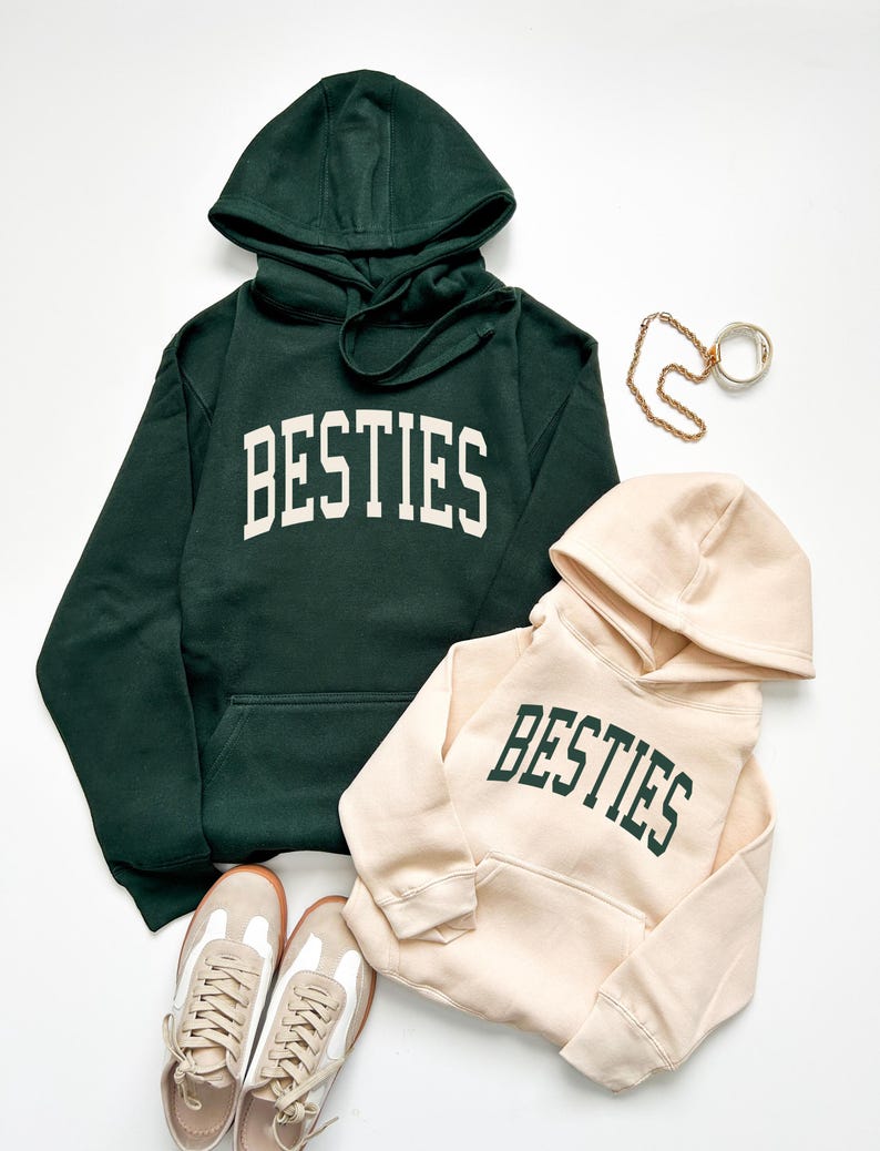 May include: Two hooded sweatshirts, one dark green and one beige, both with the word "BESTIES" in bold, white and green lettering. A pair of beige and white sneakers and gold jewelry are also in the image.
